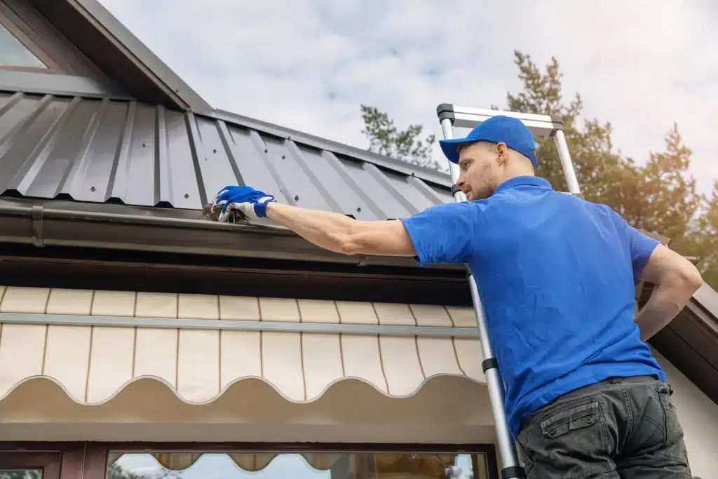 A worker on a ladder examines a metal roof and gutter, indicating re-roofing work.